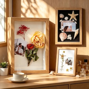 Op de afbeelding: Drie licht houten schaduwboxlijsten met gedroogde bloemen, schelpen en foto's. De grootste lijst bevat een gele roos en rode bloemen, de lijst rechtsboven toont een zeester en schelpen, en de onderste lijst bevat gedroogde wilde bloemen.