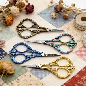 Puede incluir: Cuatro pares de tijeras de bordado decorativas en bronce, iridiscente, azul y dorado. Cada par presenta mangos ornamentados con diseños florales y los nombres "Amelin", "Sophia", "Elena" y "Jamie" grabados en las hojas. Una cinta métrica y flores secas en el fondo.