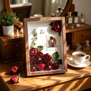 Könnte beinhalten: Ein rechteckiger hölzerner Schattenkastenrahmen mit getrockneten roten Rosen, grünen Blättern, weißen Perlen und einem kleinen Foto eines Paares. Der Rahmen trägt oben den eingravierten Text 01.16.2025 und unten The First Flowers, The Beginning of Our Love.