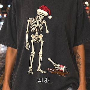 May include: A charcoal gray t-shirt featuring a graphic of a skeleton wearing a red and white Santa hat. The skeleton stands over a spilled bottle labeled Peppermint Mocha and a candy cane. The text Well Shit... is printed below the graphic.