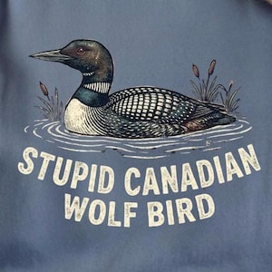 May include: A blue t-shirt with a graphic of a loon in water, and the text "STUPID CANADIAN WOLF BIRD". The bird is depicted with black, white, and grey markings. The shirt has short sleeves and a relaxed fit, suitable for casual wear.