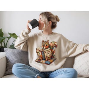Peut inclure: Sweat-shirt beige avec un écureuil de dessin animé lisant un livre intitulé "Acorn Tales". L'écureuil est assis sur un coussin patchwork. Le sweat-shirt est de couleur neutre, adapté au port décontracté.