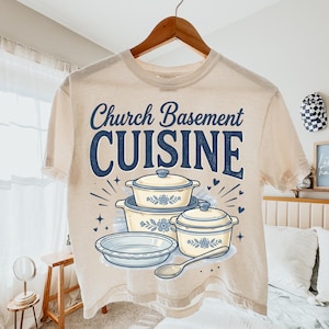 May include: Cream-colored t-shirt with the text "Church Basement CUISINE" in navy blue. The design features illustrations of casserole dishes and a spoon, with floral accents. The shirt is hanging on a wooden hanger.
