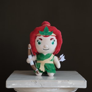 May include: A plush doll of a female archer, standing on a white pedestal. The doll has red hair, a green outfit, and holds a bow and arrow. The doll's face is detailed with large eyes and a gentle smile. The background is a dark brown.