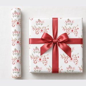 May include: White wrapping paper featuring a repeating pattern of baby clothes and apples hanging on a clothesline with the text "the APPLE of our eye" in red. A roll of the paper sits next to a square gift box wrapped in the paper with a red ribbon.