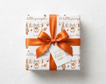May include: A square gift box wrapped in white paper featuring a repeating pattern of baby clothes on a clothesline, pumpkins, and autumn leaves. The text reads little pumpkin is on the way. A burnt orange satin ribbon bow and a tag reading FOR THE little one.