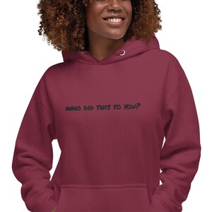 May include: A maroon hoodie with a drawstring hood and a front pocket. The text "WHO DID THIS TO YOU?" is printed in green across the chest. The person in the image is smiling.