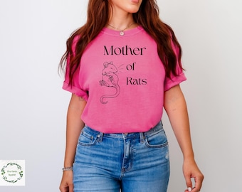 May include: A bright pink short-sleeved t-shirt featuring a black line-art illustration of a rat and the text "Mother of Rats" printed in a serif font on the chest. The shirt is paired with blue denim jeans.