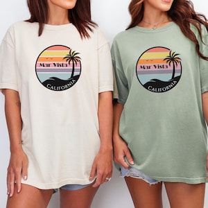 May include: Two oversized t-shirts, one cream and one sage green, each featuring a circular graphic. The graphic includes a sunset, palm tree, and the words "Mar Vista California". The shirts are casual and comfortable.