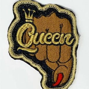 May include: Embroidered patch featuring a brown fist with the word "Queen" in gold script. A gold crown sits above the word. The patch has a black border with gold stitching and a red accent.