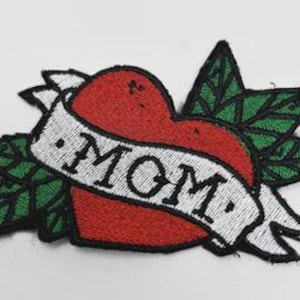 May include: Embroidered patch featuring a red heart with the word "MOM" on a white banner. The heart is surrounded by green leaves with black outlines. The patch has a black border and is likely for clothing or accessories.
