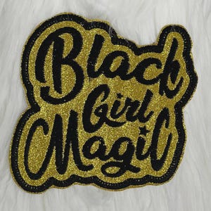 May include: A gold glitter patch with the words "Black Girl Magic" in black script. The patch has a black border and is set against a white, fluffy background. The design is bold and eye-catching.