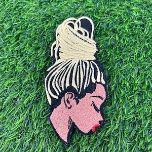 May include: Embroidered patch featuring a side profile of a woman with her eyes closed and hair styled in a bun. The patch has a black outline, with blonde hair, a pink face, and red lips. The patch is on a green, grassy surface.