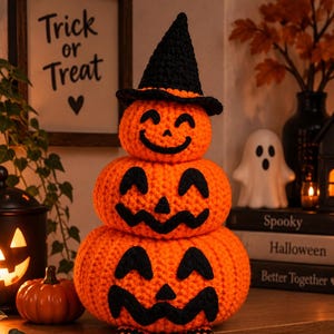 Puede incluir: Una pila de tres calabazas naranjas tejidas a ganchillo con caras negras cosidas y un sombrero puntiagudo negro. La calabaza inferior tiene piernas largas y colgantes con rayas horizontales naranjas y negras y pies negros decorativos.