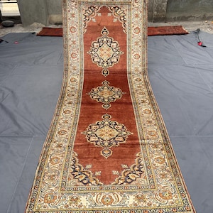 May include: A long, rectangular rug with an intricate floral pattern in shades of red, cream, and navy blue. The rug features a central medallion design and a detailed border with fringe on both ends. The rug is displayed on a gray surface.