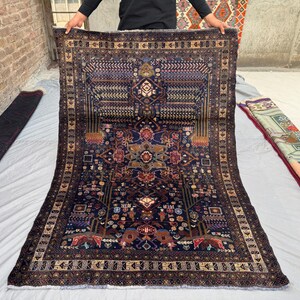 May include: A rectangular, dark blue rug with an intricate, symmetrical pattern. The rug features a central floral design with red, brown, and gold accents, surrounded by a detailed border. The rug is displayed on a light gray surface.