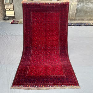 May include: A rectangular, red rug with a detailed geometric pattern. The rug has a dark red border and a lighter red center, with a fringe along the bottom edge. The rug is displayed on a light gray surface.