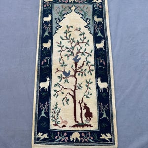 May include: Rectangular rug with a cream-colored central panel featuring a tree with birds and a rabbit. The border is dark blue with white sheep and floral designs. The rug has a cream fringe.