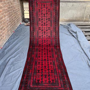Może przedstawiać: Długi, prostokątny czerwony dywan z powtarzającym się geometrycznym wzorem w kolorze czarnym. Dywan ma obramowanie i frędzle na końcach. Dywan jest trzymany przez osobę.