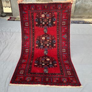 May include: A rectangular red rug with a central design of three stylized floral arrangements in black, blue, and white. The rug has a detailed border with repeating patterns and a fringed edge. The rug is displayed against a neutral background.