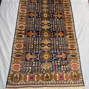 Puede incluir: Una alfombra rectangular, tejida a mano, con un fondo azul oscuro y intrincados patrones geométricos en tonos marrones, dorados, verdes y rojos. La alfombra presenta un diseño de medallón central y un borde detallado con flecos.