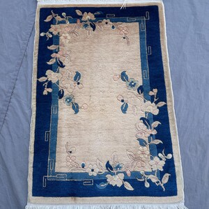 Può includere: Piccolo tappeto rettangolare con un centro beige e un bordo blu scuro. Il bordo presenta un motivo floreale con fiori bianchi e rosa e foglie blu. Il tappeto ha una frangia alle estremità corte.