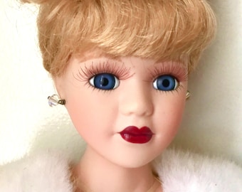 Puede incluir: Muñeca de porcelana con cabello rubio recogido en un moño, ojos azules y labios rojos. La muñeca lleva una estola blanca de piel sintética y pendientes pequeños. Un adorno para la cabeza decorativo con perlas y cristales adorna el cabello de la muñeca.