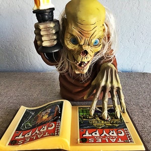 May include: A grotesque, yellow-skinned crypt keeper figure with blue eyes and white hair, holding a candle and reaching towards an open book. The book displays the text "Tales from the Crypt" on its pages, with colorful illustrations.