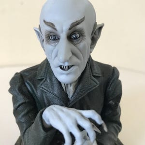 Pode incluir: Estatueta de um vampiro careca de pele pálida, com orelhas pontudas e dentes afiados. O vampiro usa um casaco preto e tem dedos longos e finos. A estatueta é provavelmente feita de resina ou material semelhante.