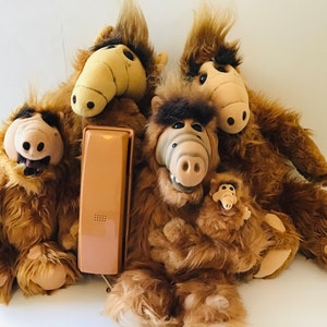 Op de afbeelding: Een verzameling pluche ALF-poppen met bruine vacht en beige gezichten. E&eacute;n pop houdt een kleinere ALF-pop vast. Een vintage beige telefoonhoorn ligt voor de poppen.