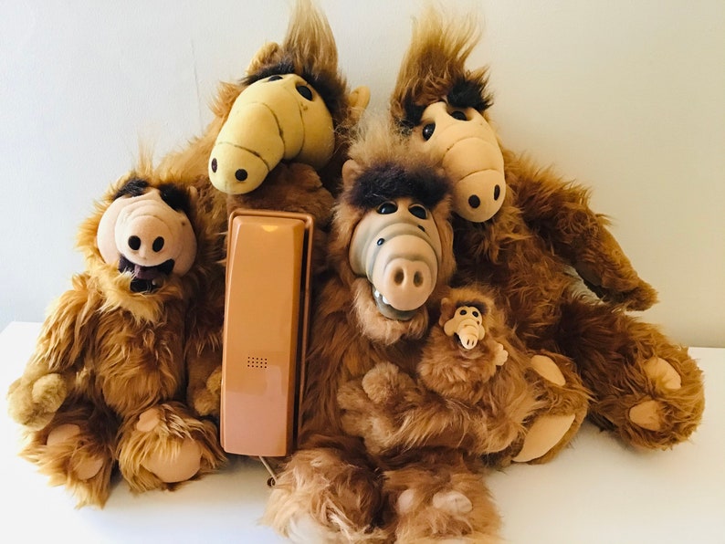 Op de afbeelding: Een verzameling pluche ALF-poppen met bruine vacht en beige gezichten. E&eacute;n pop houdt een kleinere ALF-pop vast. Een vintage beige telefoonhoorn ligt voor de poppen.