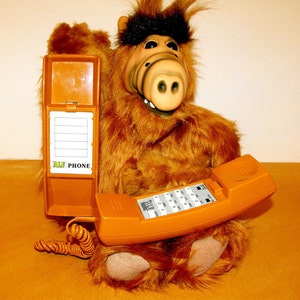 Op de afbeelding: Een bruine pluche ALF-pop met een oranje telefoon. De telefoon heeft een toetsenbord en een opgerold snoer. Een bijpassende oranje telefoongids staat open naast de pop. De tekst "ALF PHONE" is zichtbaar.
