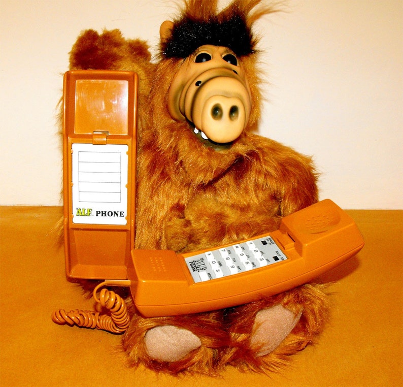 Op de afbeelding: Een bruine pluche ALF-pop met een oranje telefoon. De telefoon heeft een toetsenbord en een opgerold snoer. Een bijpassende oranje telefoongids staat open naast de pop. De tekst "ALF PHONE" is zichtbaar.
