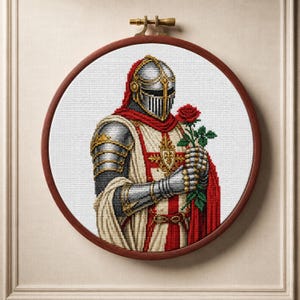 May include: A circular wooden embroidery hoop containing a cross-stitch pattern of a medieval knight in silver armour and a white tunic with a red cross. The knight holds a single red rose with green leaves and wears a helmet with a visor.