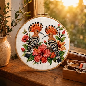 May include: A circular wooden embroidery hoop containing a cross-stitch pattern of two hoopoe birds with orange crests and black and white striped wings, surrounded by vibrant red and orange hibiscus flowers and green leaves on white fabric.