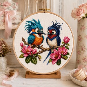 Peut inclure: Un tambour à broder circulaire en bois contenant un motif au point de croix représentant deux oiseaux à crêtes bleues hérissées perchés sur une branche brune. Les oiseaux sont entourés de fleurs roses et de feuilles vertes sur fond blanc.