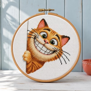 Könnte beinhalten: Ein runder Stickrahmen aus Holz mit einem Kreuzstichmuster eines orangefarbenen getigerten Katers mit großen grünen Augen und einem breiten, zahnigen Grinsen. Die Katze schaut hinter einer vertikalen Kante hervor und zeigt eine Pfote.