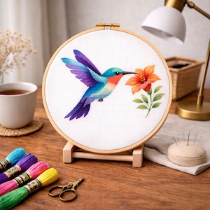 May include: A colorful hummingbird cross-stitch embroidery in a wooden hoop. The bird has blue, purple, and teal feathers, and is feeding from an orange flower. The embroidery is displayed on a wooden stand.