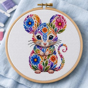 Könnte beinhalten: Ein runder Stickrahmen aus Holz mit einem Kreuzstichmuster einer Maus. Die Maus ist mit leuchtend bunten Blumen- und Blattmotiven in Blau-, Rosa-, Orange- und Grüntönen verziert, die auf einem weißen Stoffhintergrund gestickt sind.