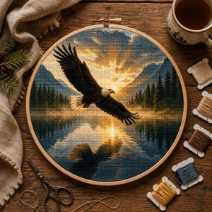 May include: A circular wooden embroidery hoop containing a cross-stitch project of a bald eagle in flight over a lake at sunset. The scene features dark blue mountains, evergreen trees, and a bright golden sun reflecting on the water surface.