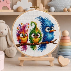 Puede incluir: Un bordado de punto de cruz colorido con tres pájaros caprichosos posados en una rama. Los pájaros son de color naranja, verde y azul, con detalles de plumas. El bordado se muestra en un bastidor de madera sobre un caballete pequeño.