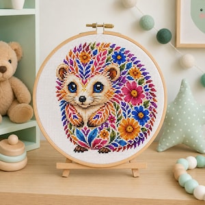 May include: A circular wooden embroidery hoop containing a cross-stitch pattern of a hedgehog. The hedgehog is decorated with vibrant, multi-colored floral and leaf motifs in shades of pink, blue, orange, and purple, set against a white fabric.