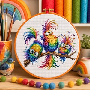 May include: A circular embroidery hoop containing a cross-stitch pattern of three colorful, whimsical birds with large eyes and spiky feathers perched on a brown branch. The birds feature a vibrant rainbow gradient of colors against white fabric.