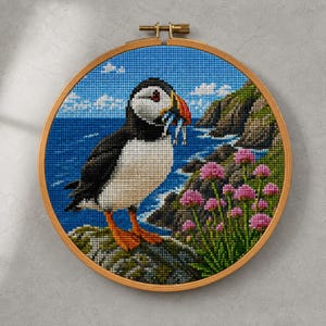 May include: A circular wooden embroidery hoop containing a cross-stitch pattern of a puffin standing on a rocky cliffside. The puffin has black and white feathers, an orange beak holding small fish, and orange feet, set against a blue ocean background.