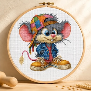 Puede incluir: Un bordado de punto de cruz de un ratón de dibujos animados que lleva una gorra de béisbol de patchwork multicolor, una chaqueta vaquera azul y zapatillas amarillas. El ratón tiene grandes orejas rosas, ojos azules y una cola larga y fina, todo enmarcado en un bastidor de madera redondo.