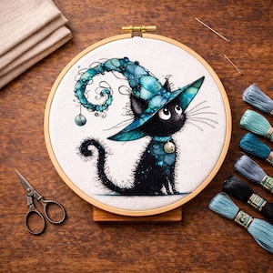 May include: A circular wooden embroidery hoop containing white fabric with a cross-stitch design of a black cat wearing a large, swirling teal and blue pointed hat. The cat has large eyes and a small bell collar. Embroidery floss and scissors are nearby.