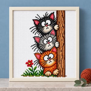 Op de afbeelding: Een ingelijst kruissteek borduurwerk met drie cartoonkatten die verticaal tegen een bruine boomstam zijn gestapeld. De bovenste kat is zwart, de middelste grijs en de onderste oranje. Een kleine rode bloem groeit in het groene gras onderaan.