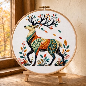 Peut inclure: Une broderie au point de croix représentant un cerf aux grands bois, orné d'un motif coloré de feuilles et de formes géométriques dans des tons orange, bleu sarcelle et jaune. L'ouvrage est présenté dans un tambour en bois sur un support.