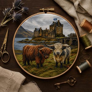 May include: A circular wooden embroidery hoop containing a cross-stitch project depicting two Highland cattle standing in a grassy field before a stone castle on a lake. The castle features a small blue and white flag on its highest tower.
