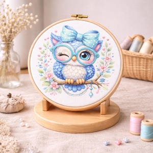 Puede incluir: Un bastidor de bordado circular de madera que contiene un patrón de punto de cruz de un búho azul con gafas grandes de color azul claro y un lazo a juego con lunares. El búho está posado sobre una rama rodeada de flores y hojas.